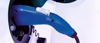 A blue electric vehicle charging plug connected to a white electric car's charging port. The close-up image highlights the handle and connector of the charging plug.