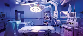 A modern operating room filled with medical equipment. There's a surgical table in the center, surrounded by monitors, overhead lights, and various machines. The lighting is dim with a bluish hue, giving an ambient atmosphere.