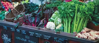 A market stall displays a variety of fresh vegetables, including leafy greens, cauliflower, celery, and parsnips. Chalkboard signs state "Home Grown Produce" and "No Chemicals Used." Various fruits and vegetables are neatly arranged in the background.