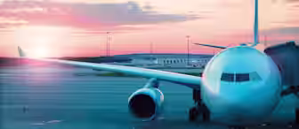 A large passenger airplane is parked at an airport gate during sunset. The sky features a mix of pink, orange, and blue hues while the sun is partially visible near the horizon. The airplane is connected to the gate by a jet bridge.