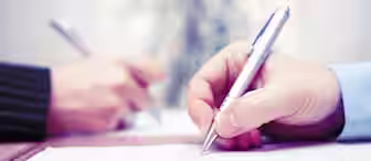 Close-up of two people writing on documents with silver pens. The focus is on their hands, one person wearing a suit jacket and the other in a light blue shirt, both engaged in what appears to be a formal signing or writing activity.