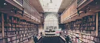 A cozy, two-story library with wooden bookshelves lining both sides and a central staircase leading to the upper level. The floor is wooden, and natural light pours in from a large, arched window in the background, illuminating the spacious interior.