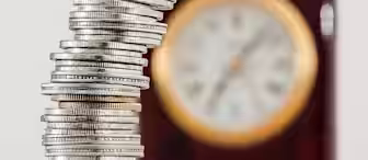 A tall, precarious stack of silver coins is in the foreground, with some coins scattered at the base. In the blurred background, there's a round clock with a gold-colored rim, indicating the passage of time related to the accumulation of wealth.
