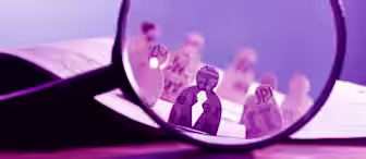 A large magnifying glass focuses on paper cut-outs of human figures, each decorated with various letters and text passages. The background shows more paper figures out of focus, positioned on an open book, creating a scene with text and paper characters.