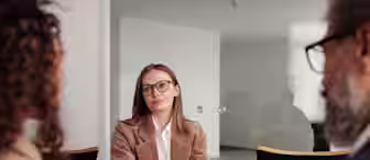 A woman wearing glasses and a brown blazer sits at a table with her hands folded, facing two blurred individuals in an office setting. She appears engaged and attentive, with a glass door and office furnishings in the background.