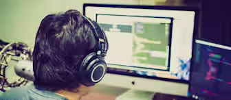 A person wearing headphones sits at a desk facing a large monitor that displays coding scripts. Another smaller monitor to the right also shows code. Various electronic devices and objects are on the desk. The scene suggests a focused work environment.