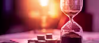 A close-up of an hourglass with sand flowing into the lower chamber, placed on a table scattered with coins and a few stacks of coins. The background is blurred with a warm, glowing light, creating a soft and nostalgic ambiance.