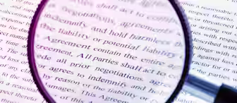 A magnifying glass held over a document with dense text, highlighting words related to agreements, negotiations, and indemnity. The view through the lens is sharp, while the surrounding text is blurred. The image emphasizes the importance of scrutinizing legal documents.