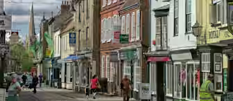A quaint street lined with colorful, historic buildings housing various shops. Pedestrians walk along the cobblestone sidewalk, some browsing store windows. Lamp posts, shop signs, and a distant church spire are visible under a partly cloudy sky.