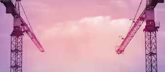 Two tall construction cranes are silhouetted against a vibrant, multi-colored sky with hues of pink, purple, and blue. The cranes face each other, forming a symmetrical frame, while wispy clouds drift in the background.