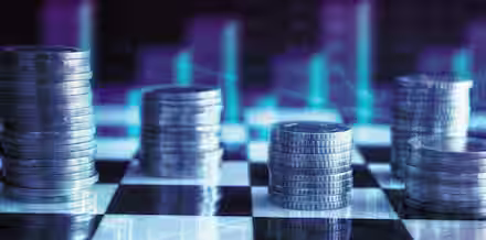 Stacks of coins are arranged in rows on a black and white checkered surface. In the background, blurred bar charts and line graphs are visible, creating a financial or economic theme. The image is filled with blue and purple hues, giving it a futuristic feel.