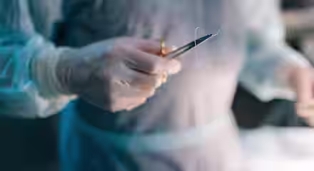 A person wearing surgical gloves and a gown holds a needle holder with a curved needle and suture thread, preparing for a medical procedure in an operating room.