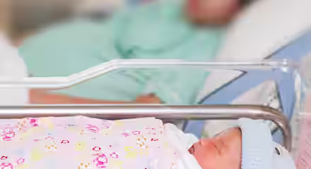 A newborn baby wearing a blue hat sleeps in a hospital bassinet, wrapped in a colorful blanket. In the blurred background, an adult lies in a hospital bed.