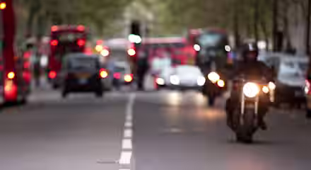 A city street scene with blurred red double-decker buses, cars, and motorcycles in the background. The road is lined with trees, and a motorcyclist rides toward the camera on the right side, with the streetlights illuminating.