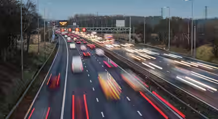A busy multi-lane highway at dusk with blurred headlights and taillights from fast-moving vehicles, bordered by grassy areas and trees, with an overhead traffic sign in the distance.