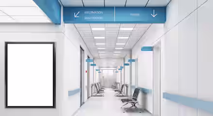 Modern hospital corridor with white walls, blue accents, empty chairs, and signs for information, registration, wards, and CT room. A large blank notice board is on the left wall. The area is clean and brightly lit.