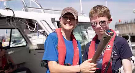 Two people wearing red life jackets and sunglasses smile on a boat. One holds a metal object while the other places a hand on their shoulder. The background shows a sunny marina with boats and a pier.