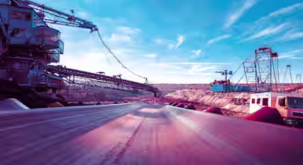 A conveyor belt runs through an open-pit mining site under a bright blue sky. Heavy machinery and cranes can be seen on either side, and rugged, rocky terrain stretches into the distance. A truck is parked to the right, emphasizing the scale of the operation.
