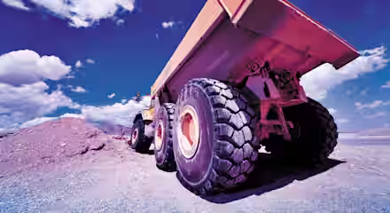 Low-angle view of a large orange dump truck with massive tires, positioned next to a pile of dirt under a vivid blue sky with scattered white clouds. The image captures the robust and heavy-duty nature of the construction vehicle.