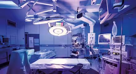 A modern operating room filled with medical equipment. There's a surgical table in the center, surrounded by monitors, overhead lights, and various machines. The lighting is dim with a bluish hue, giving an ambient atmosphere.