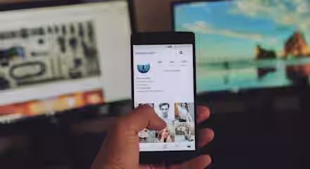 A person holds a smartphone showing an Instagram profile. In the background, there are two computer monitors; the left screen displays a technical diagram and the right screen shows a scenic beach landscape. The surroundings are dimly lit.