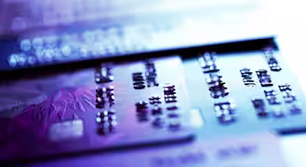 Close-up image of several credit cards laid out flat, with focus on the raised numbers of one card in the foreground. The image is illuminated with a mix of purple and blue light, giving it a modern and tech-savvy appearance.