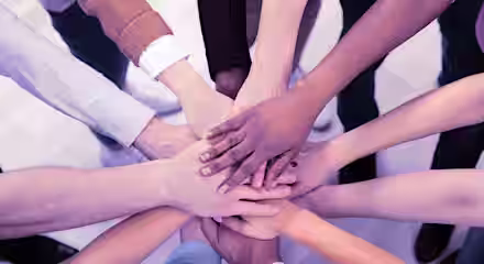 A group of diverse people standing in a circle place their hands on top of each other in the center, symbolizing unity and teamwork. The hands vary in skin tones and clothing styles, representing inclusivity and collaboration. The background is a light-colored floor.