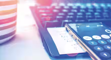 A close-up of a desk workspace with a striped mug, a computer keyboard, and a smartphone displaying a calculator app. A piece of paper with handwritten notes is beneath the phone. Sunlight creates lens flare effects on the image.