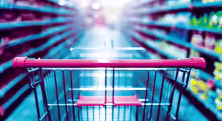 A shopping cart with a red handle is in focus in the foreground of a brightly lit supermarket aisle, surrounded by shelves stocked with a variety of products. The background is blurred, emphasizing the perspective from the shopper's point of view.