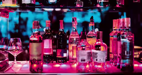 A shelf in a dimly lit bar displaying various bottles of liquor, including wine, whiskey, and vodka, illuminated by pink and blue neon lights. Two empty wine glasses are placed on the bottom shelf with a vibrant, colorful background.