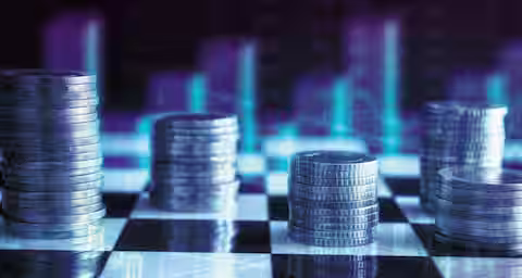 Stacks of coins are arranged in rows on a black and white checkered surface. In the background, blurred bar charts and line graphs are visible, creating a financial or economic theme. The image is filled with blue and purple hues, giving it a futuristic feel.