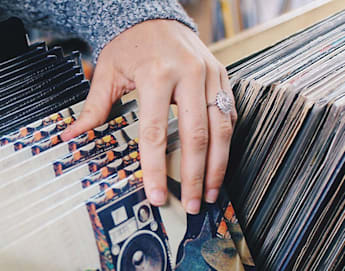 A person flips through a collection of vinyl records in a store. Some of the records are labeled with names such as "Jamie T" and "Talking Heads." The person's hands are visible, one adorned with a ring. The records are neatly organized in wooden compartments.