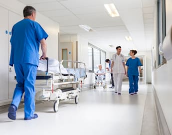 A hospital corridor with medical staff. A healthcare worker in blue scrubs pushes an empty gurney, while two other medical professionals, one in white uniform and one in blue, walk and talk. In the background, an elderly patient in a wheelchair is accompanied by a staff member.