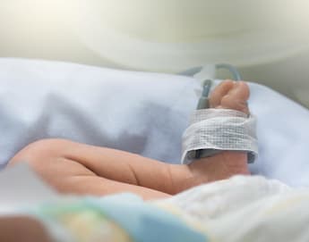 Close-up of a baby's foot with a medical sensor attached to it, lying on a white sheet in a hospital. The tiny foot has a bandage securing the sensor, indicating the baby is under medical care. Soft lighting gives a calm atmosphere to the scene.