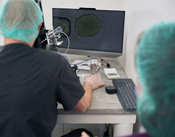 Two individuals in a laboratory setting are focused on a computer screen displaying a microscopic image. They are wearing surgical caps and gowns, indicating a sterile environment. The one on the left is manipulating a device, likely part of the microscope setup.