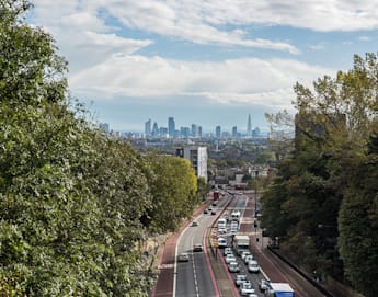 A view from an elevated position overlooking a busy road with cars and buses, flanked by trees on both sides. In the distance, the skyline of a large city with various high-rise buildings and the iconic Shard building can be seen under a partly cloudy sky.