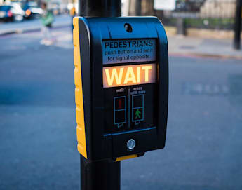 A pedestrian crossing signal box on a street. The box has a lit "WAIT" sign, indicating pedestrians should wait before crossing. The background shows a blurred view of the street and a person walking in the distance.