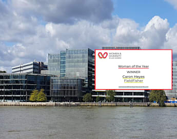 The image shows modern office buildings along the shore of a river under a partly cloudy sky. Superimposed is a graphic announcing a "Women of Diversity Award" for "Woman of the Year," awarded to Caron Heyes from Fieldfisher.