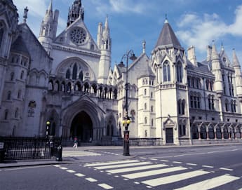 A wide view of the exterior of the Royal Courts of Justice in London on a clear day. The Gothic-style building features pointed arches, intricate stonework, and large windows. The street in front has a zebra crossing and few pedestrians.