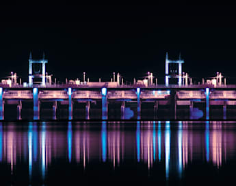 A brightly lit dam at night reflecting on the calm water below. The structure is illuminated with blue and purple lights, creating a vivid and serene scene.