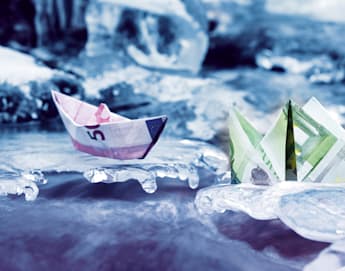 Two origami boats made from Euro banknotes are set on icy surfaces. The boat on the left is crafted from a purple €5 note, while the one on the right uses a green €100 note. The scene is surrounded by ice formations, giving a cold, frozen backdrop.