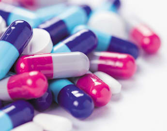 A close-up image of a pile of various pills and capsules. The capsules are predominantly in shades of blue, pink, white, and purple, while the pills are mostly white. The background is a plain, light surface.