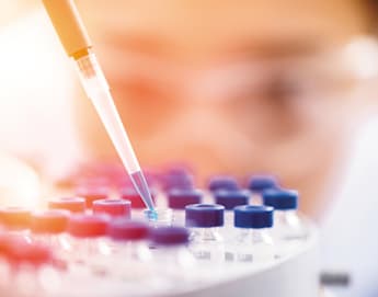 A pipette is dispensing a blue liquid into a tiny vial or test tube among an array of similar vials, each with blue caps. The background is blurred, possibly indicating a person focusing on the task, suggesting a laboratory setting.