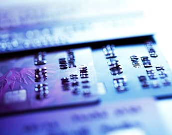 Close-up image of several credit cards laid out flat, with focus on the raised numbers of one card in the foreground. The image is illuminated with a mix of purple and blue light, giving it a modern and tech-savvy appearance.