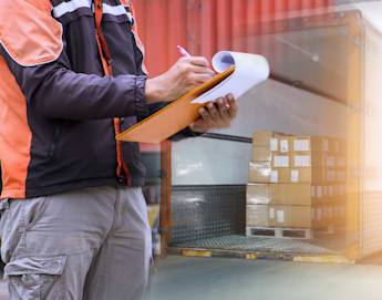 A person wearing a black and orange jacket holds a clipboard and writes notes. Behind them, a truck is being loaded with several stacks of cardboard boxes. The scene suggests logistics or delivery operations, with a focus on inventory or shipment documentation.