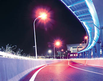 A vibrant nighttime scene of a curving, empty road illuminated by colorful neon lights in shades of blue, pink, and purple. Streetlamps with red-orange lights line the road, with a modern structure featuring similar neon lighting visible in the background.