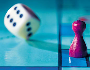 A close-up image of a red game piece and a tumbling white die with black dots on a blue game board. The die is shown mid-roll, indicating movement, while the game piece stands stationary. The game board has a grid pattern.