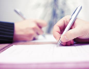 Close-up of two people writing on documents with silver pens. The focus is on their hands, one person wearing a suit jacket and the other in a light blue shirt, both engaged in what appears to be a formal signing or writing activity.