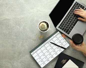 A person using a laptop with one hand and holding a coffee cup with the other. The desk also features a calendar, a small potted cactus, a pen, paper clips, and a closed notebook. The setting suggests an organized work or study environment.