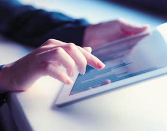 A person interacts with a tablet screen, using their index finger to touch the device. The tablet displays a chart or graph with bars, indicating data is being viewed or analyzed. The background is softly blurred, focusing attention on the hands and the tablet.